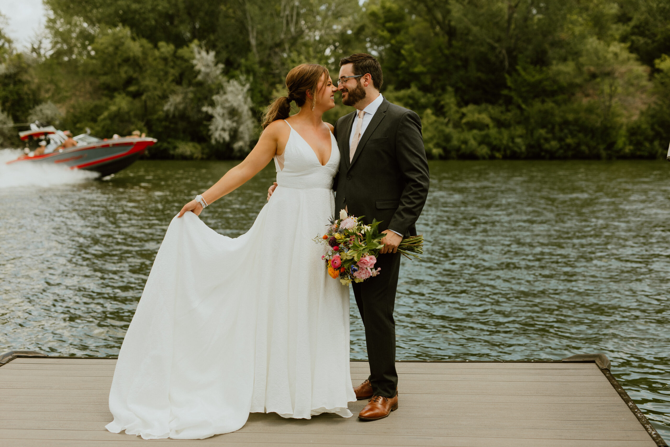 bride and groom wedding photo by lake with boat in distance at golden hour bride is holding flowers and they are touching noses